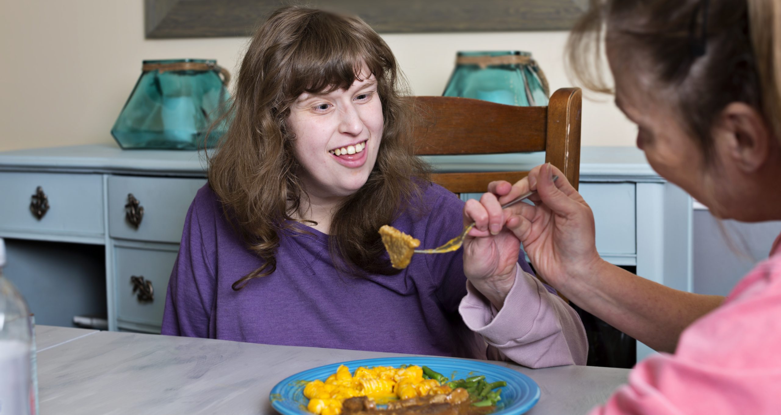 En kvinna med lila tröja får mat av en person som jobbar på en LSS-bostad.